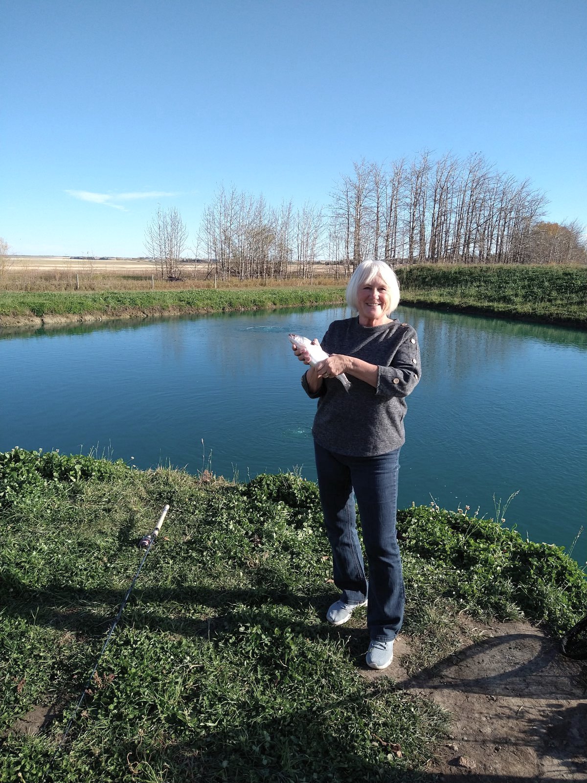 A woman smiling with her rainbow trout on the bank of the pond